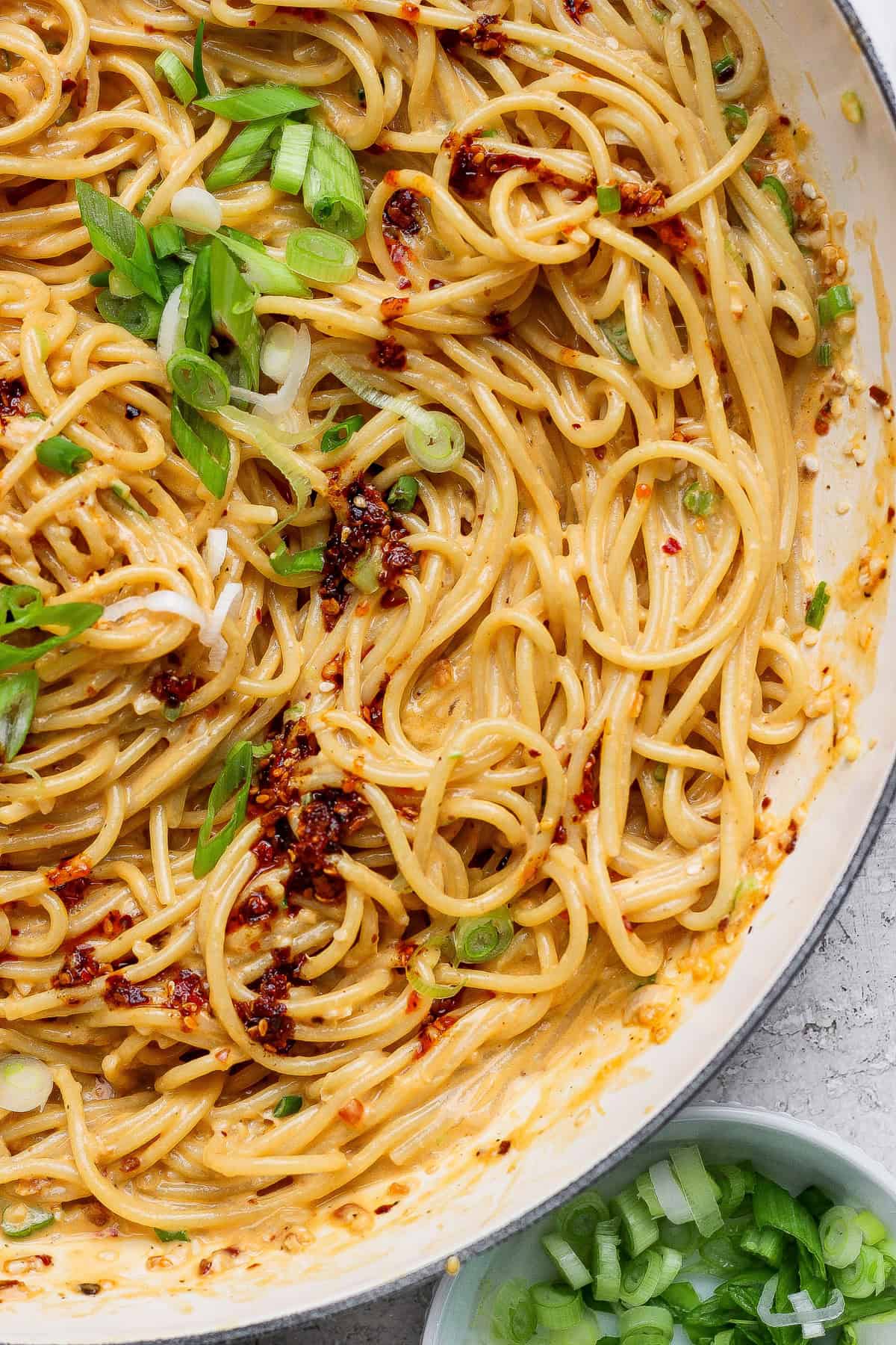 Cooked spaghetti coated in a creamy sauce, topped with chili flakes and chopped green onions, in a white pan with extra green onions in a small bowl nearby.