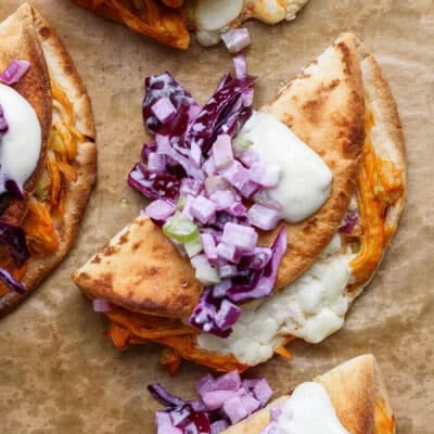 A folded quesadilla filled with melted cheese and shredded vegetables, topped with chopped red onion, purple cabbage, and a dollop of white sauce on parchment paper.