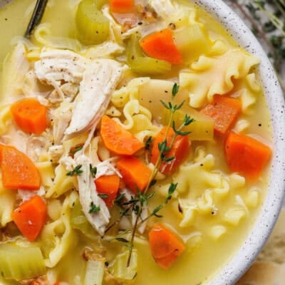 A close-up of chicken noodle soup with shredded chicken, carrots, celery, mafaldine noodles, and a sprig of thyme in a white bowl.