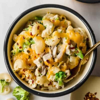 A bowl of macaroni and cheese mixed with broccoli, chicken pieces, and topped with melted cheddar cheese, with a fork inside.