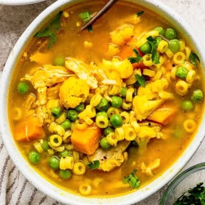 A bowl of chicken and vegetable soup with pasta, peas, cauliflower, carrots, and herbs in a clear broth.