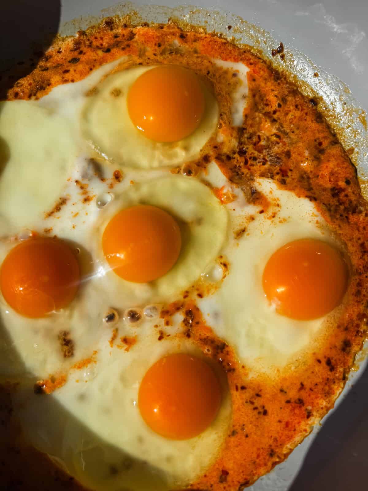 Five sunny-side-up eggs cooking in a pan, surrounded by orange-hued oil and spices.