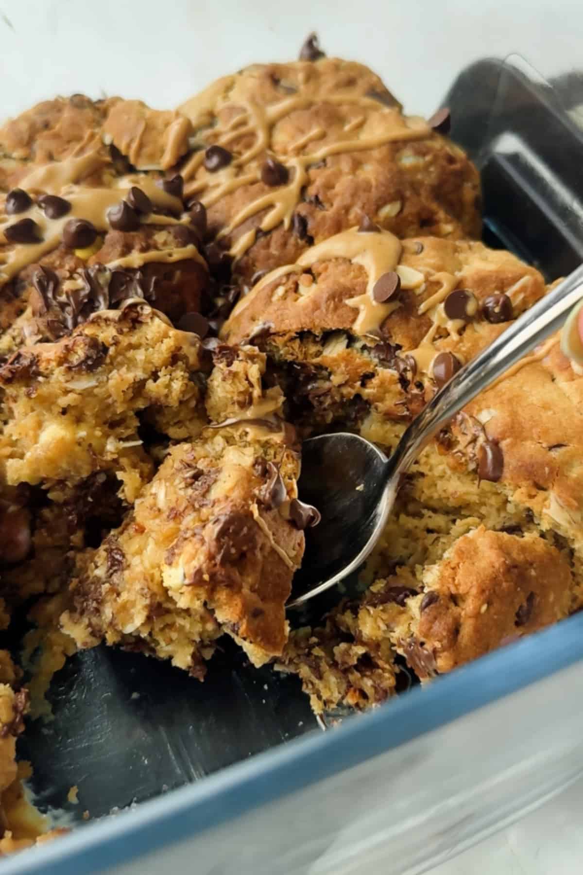 A close-up of a partially served chocolate chip cookie dessert in a glass dish, drizzled with a light glaze, with a spoon lifting a portion.