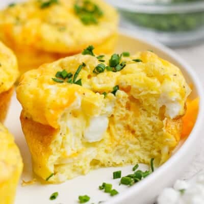 A baked egg muffin with a bite taken out, showing a fluffy texture and cheese inside, topped with chopped green onions on a white plate.