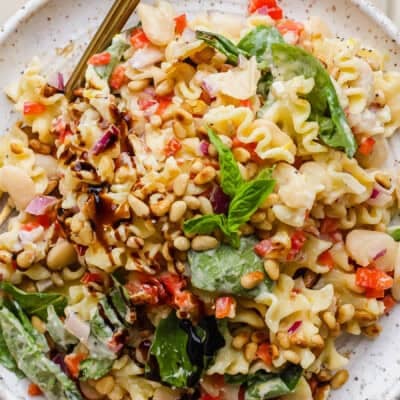 A plate of pasta salad with spinach, pine nuts, white beans, chopped red onions, red bell peppers, and a balsamic drizzle, garnished with fresh basil.