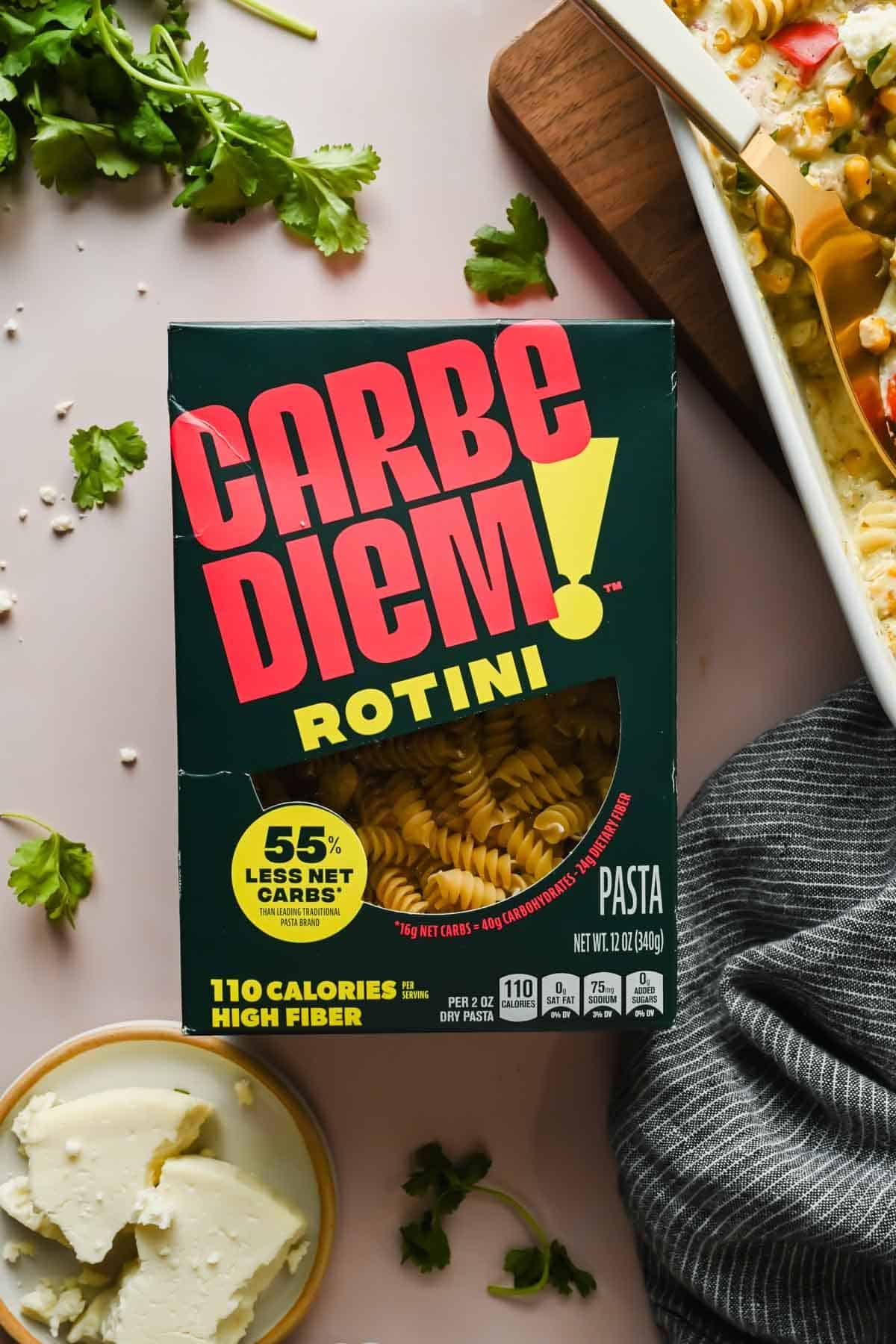 A box of Carbe Diem Rotini pasta sits on a table next to a block of cheese, fresh parsley, and a dish of baked pasta.
