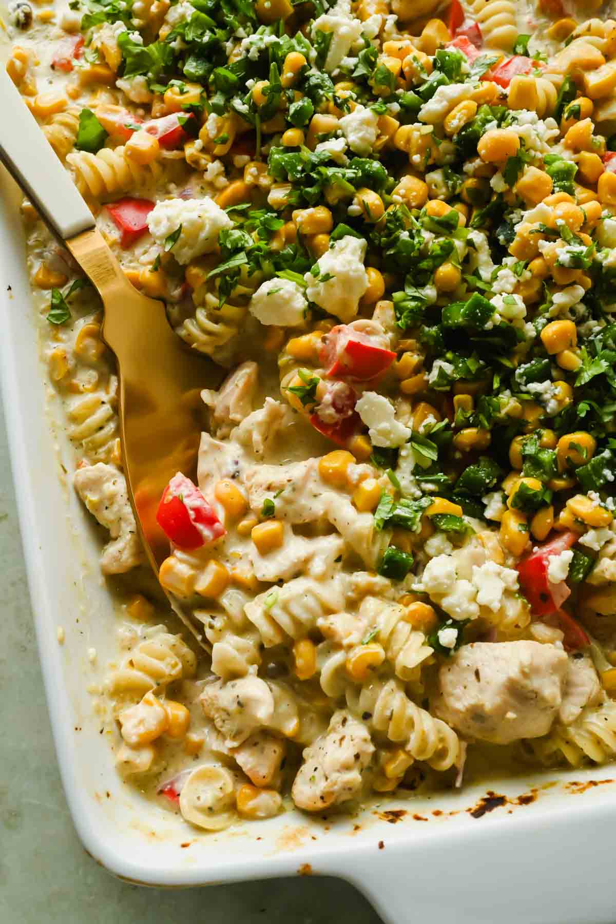 Baked pasta casserole with rotini, diced chicken, corn, red bell peppers, creamy sauce, and fresh chopped herbs, served in a white baking dish with a gold serving spoon.