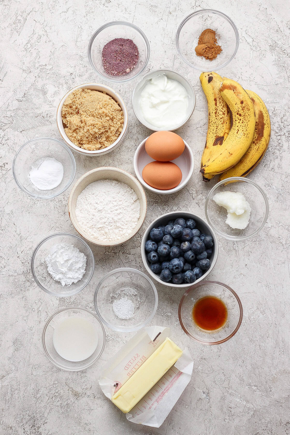 Various baking ingredients in bowls, including flour, brown sugar, eggs, ripe bananas, blueberries, butter, yogurt, vanilla extract, spices, baking powder, baking soda, and coconut oil.