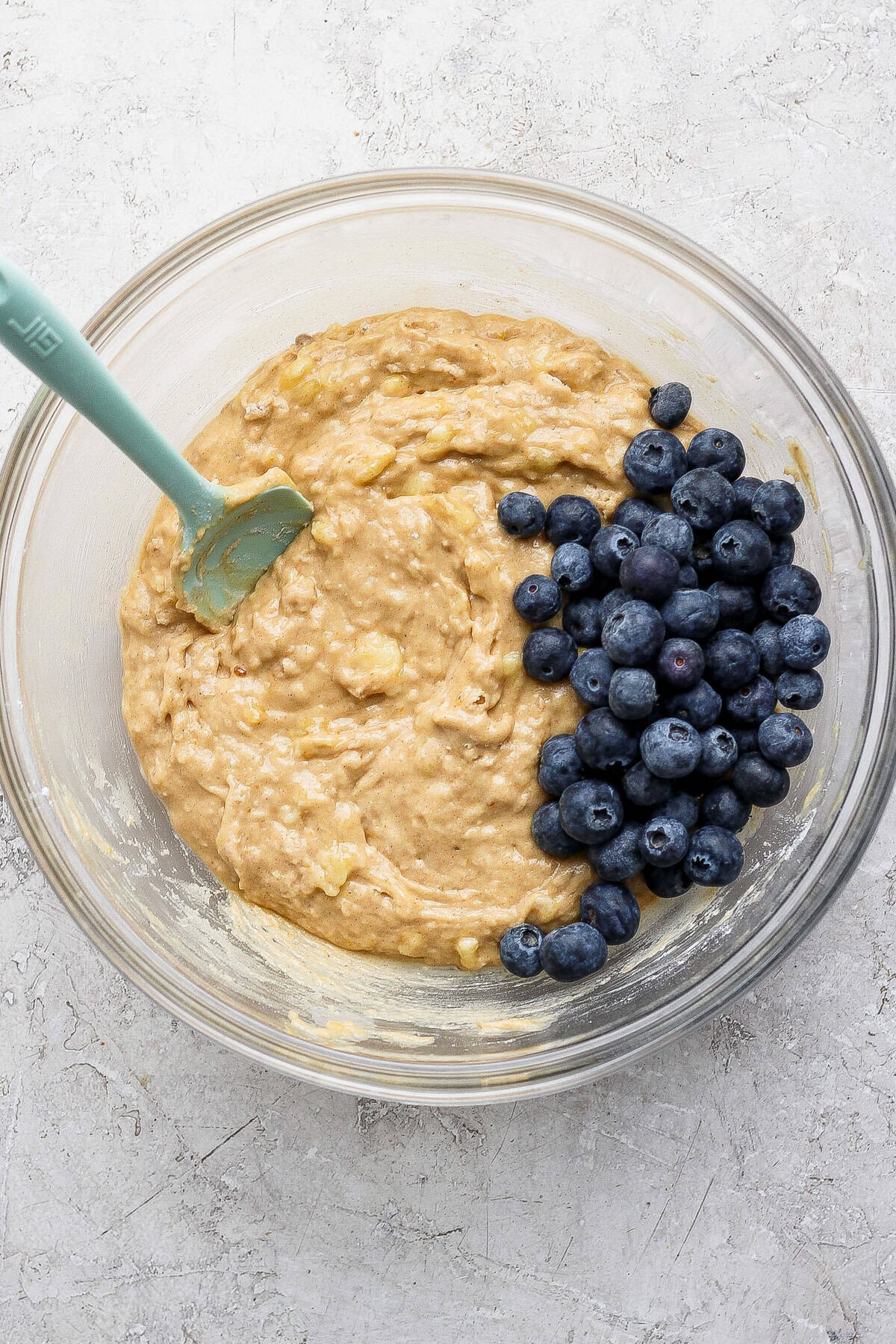A glass bowl with muffin batter and fresh blueberries, being mixed with a green spatula on a light textured surface.