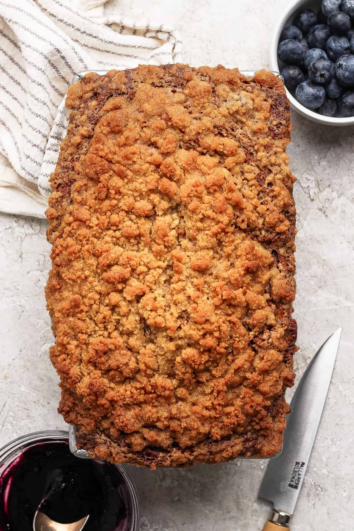 A loaf of crumb-topped bread sits on a light surface next to a knife, a bowl of blueberries, a striped cloth, and a small bowl of dark fruit jam.