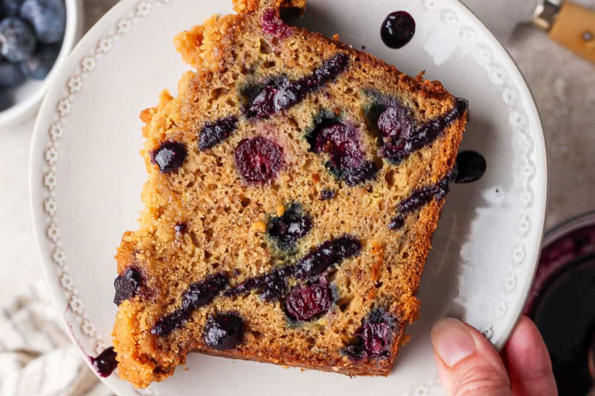 A hand holds a slice of blueberry bread on a white plate, showing blueberries and swirls of blueberry sauce throughout the slice.