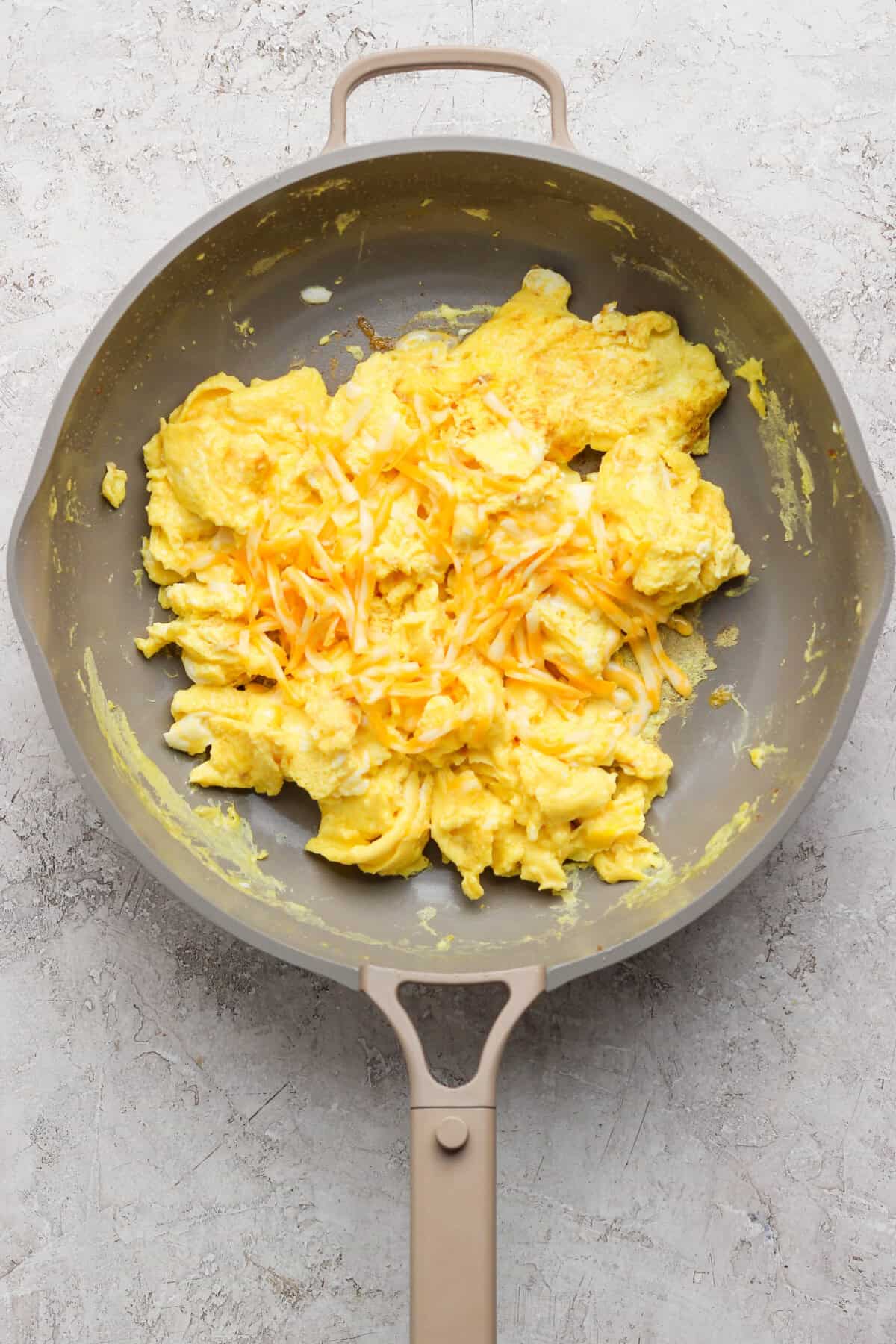 Scrambled eggs topped with shredded cheese in a nonstick skillet on a light textured surface.