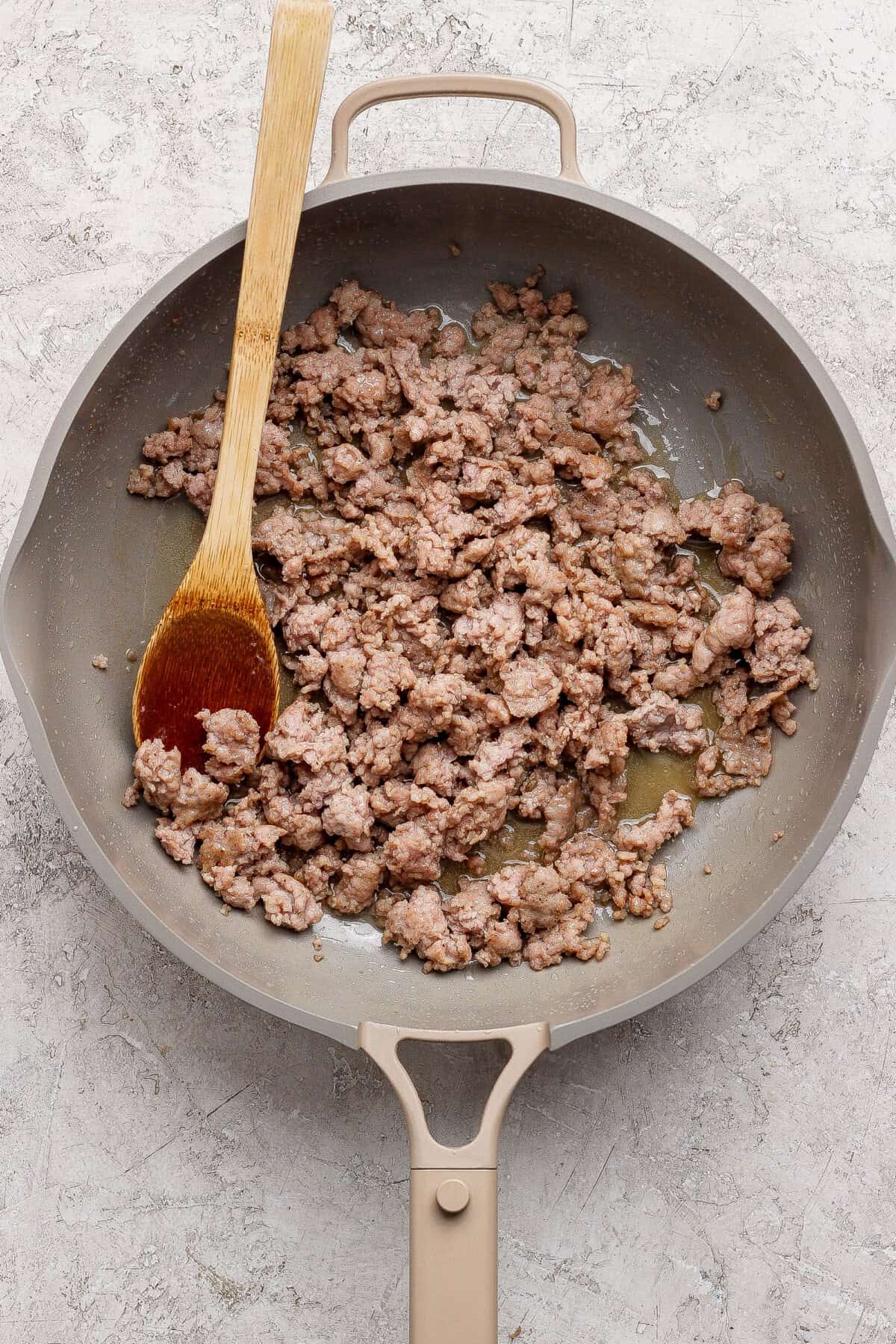 Cooked ground beef in a beige skillet with a wooden spoon, resting on a light textured surface.