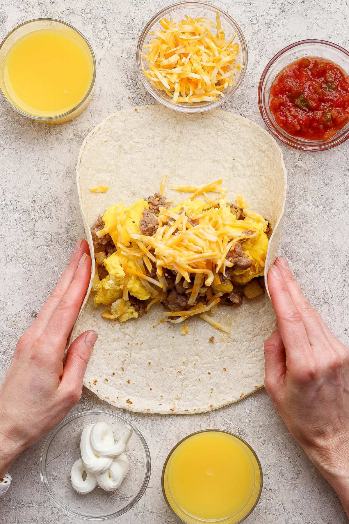 Two hands roll a tortilla filled with scrambled eggs, cheese, and meat, surrounded by bowls of shredded cheese, salsa, orange juice, and sliced onions on a light surface.
