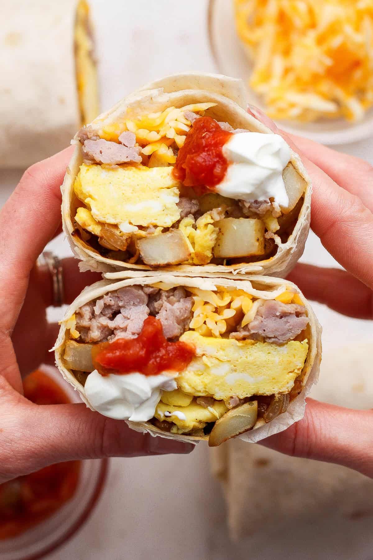 Two hands hold a halved breakfast burrito filled with scrambled eggs, sausage, potatoes, shredded cheese, salsa, and a dollop of sour cream.