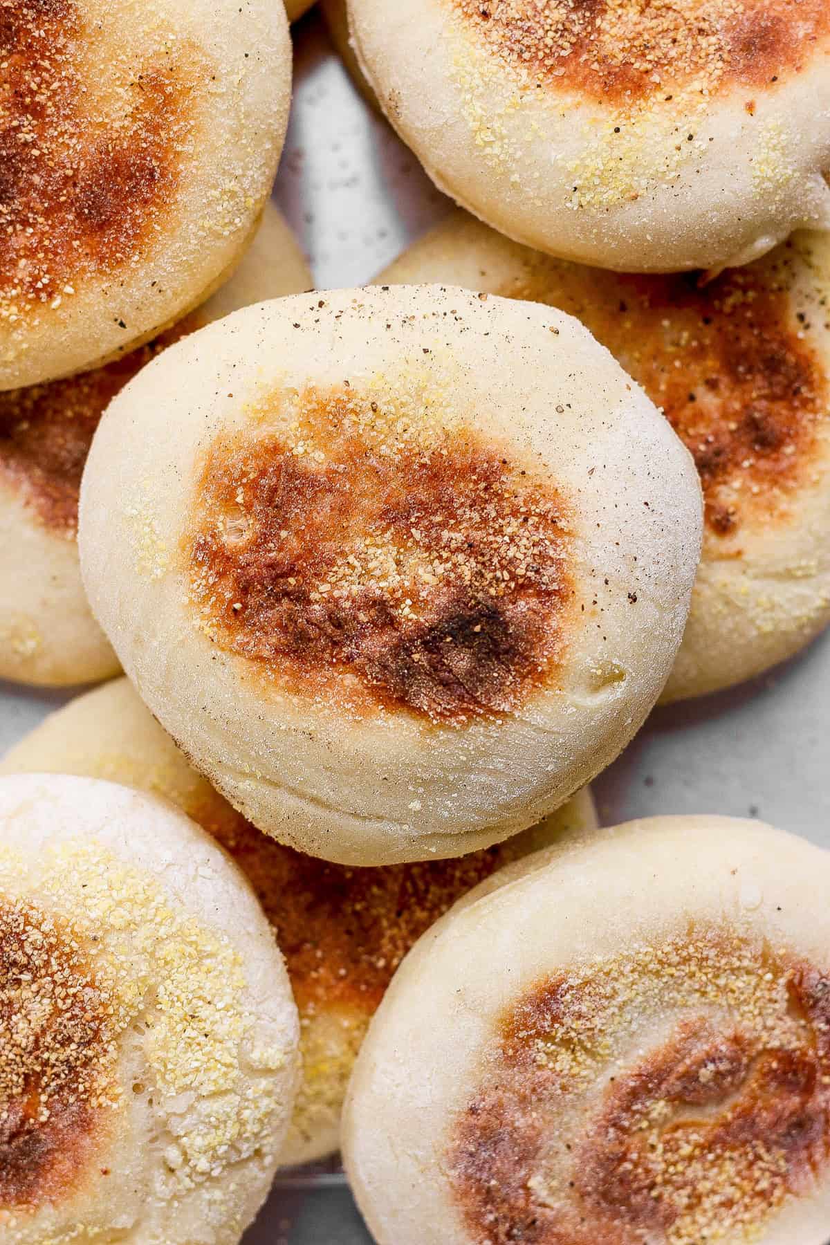 A close-up of several plain English muffins, lightly browned on top, stacked together with visible cornmeal on their surfaces.