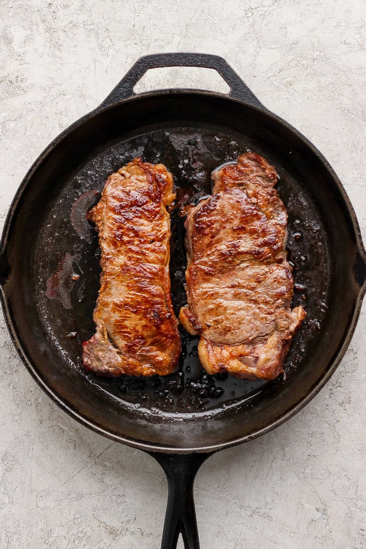 Two seared steaks are shown cooking in a black cast iron skillet on a light textured surface.
