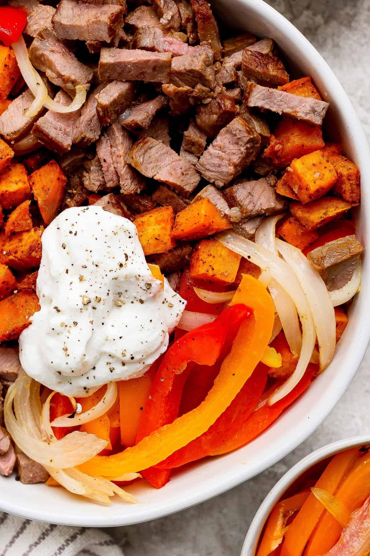 A bowl containing sliced steak, roasted sweet potatoes, saut&eacute;ed onions, red and yellow bell peppers, and a dollop of sour cream, topped with cracked black pepper.