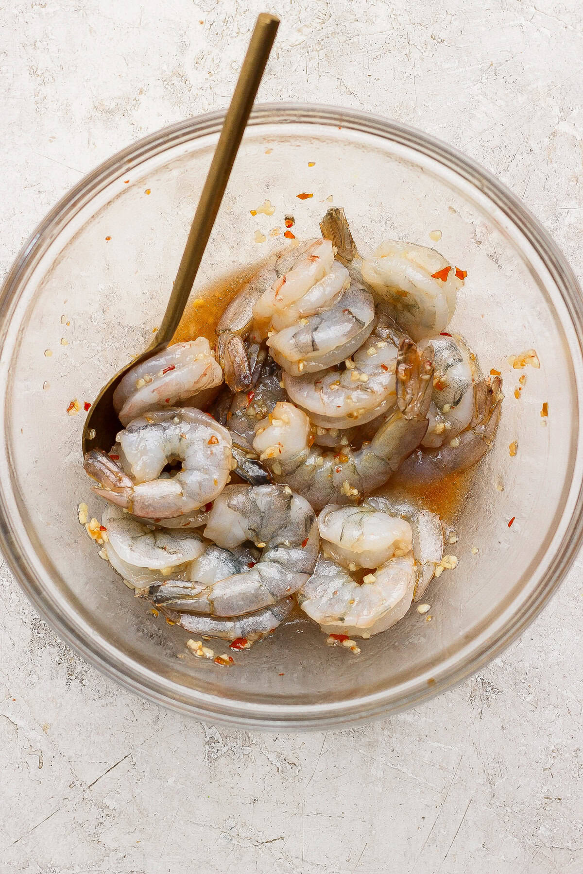 Raw shrimp marinating in a clear glass bowl with a metal spoon, mixed with a sauce containing red pepper flakes and minced garlic, on a light textured surface.