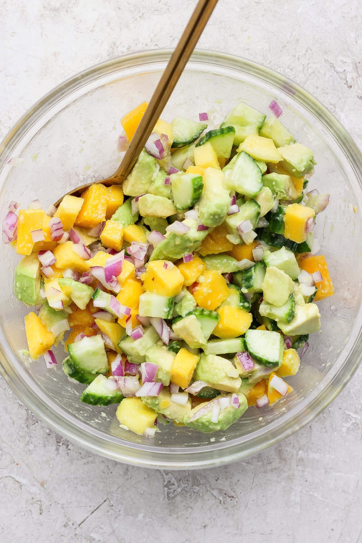 A glass bowl filled with chopped avocado, mango, cucumber, and red onion, mixed with a spoon on a light textured surface.