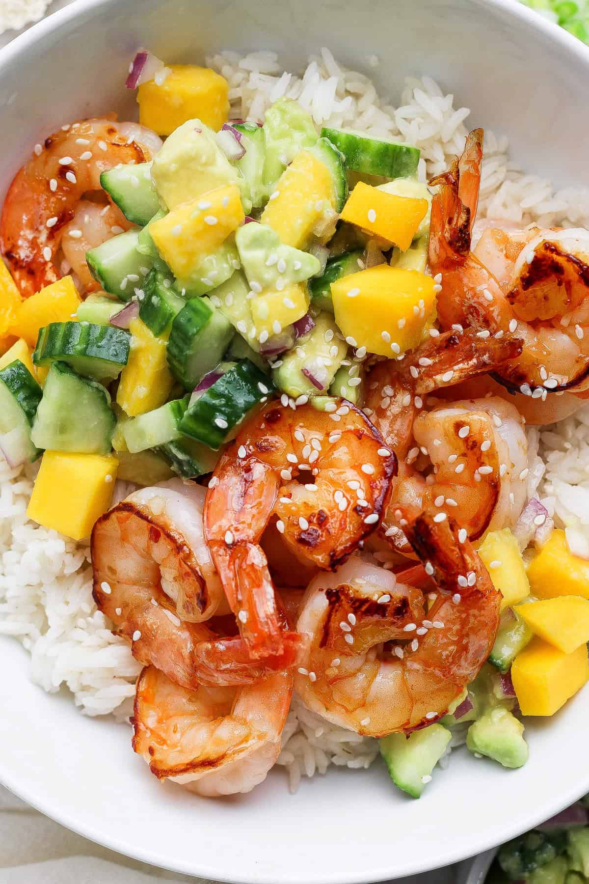A bowl of white rice topped with cooked shrimp, diced mango, avocado, cucumber, red onion, and sprinkled with sesame seeds.