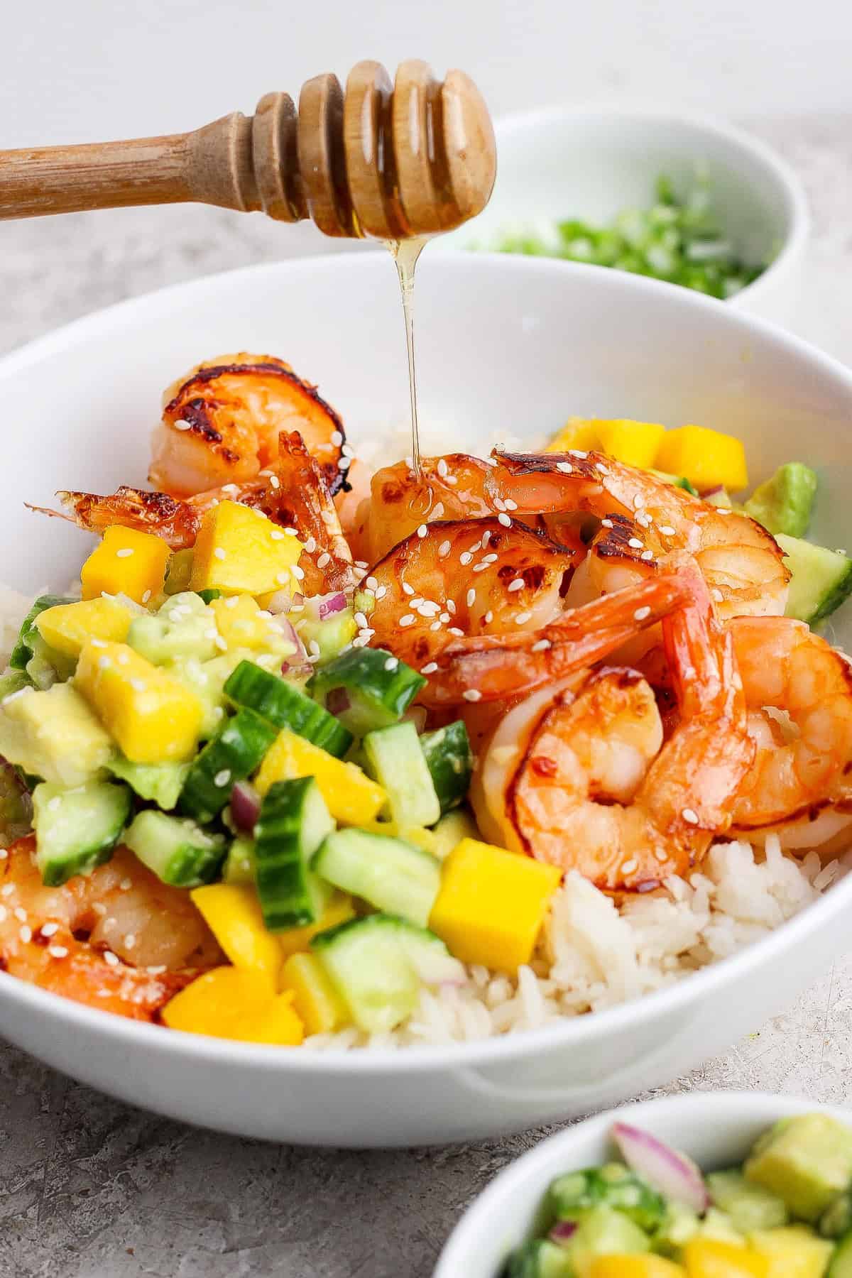 A bowl of rice topped with grilled shrimp, diced mango, cucumber, avocado, and sesame seeds, with honey being drizzled on top.