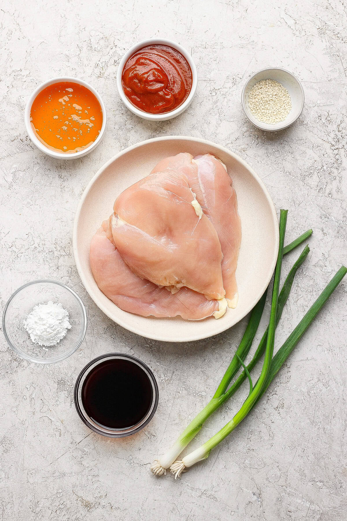 Raw chicken breasts on a plate surrounded by bowls of orange sauce, red sauce, sesame seeds, cornstarch, soy sauce, and two green onions on a light surface.