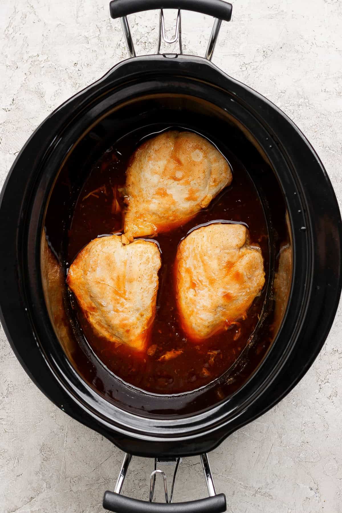Three pieces of chicken breast cooking in a slow cooker with a dark brown sauce.