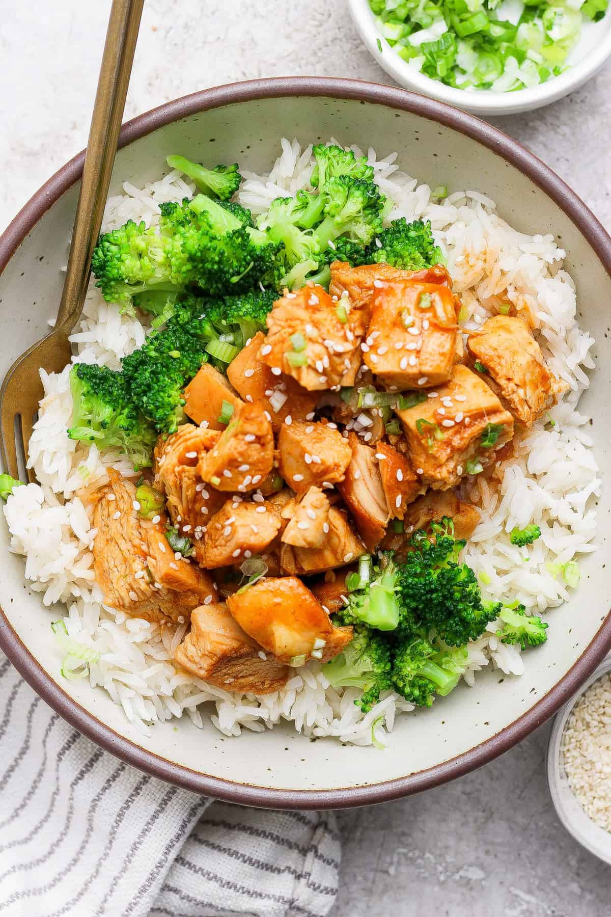 A bowl of white rice topped with broccoli and chunks of sauced chicken, garnished with sesame seeds, with a fork resting in the bowl.