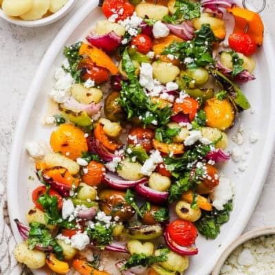 A white platter with gnocchi, roasted cherry tomatoes, bell peppers, red onions, fresh greens, and crumbled feta cheese.