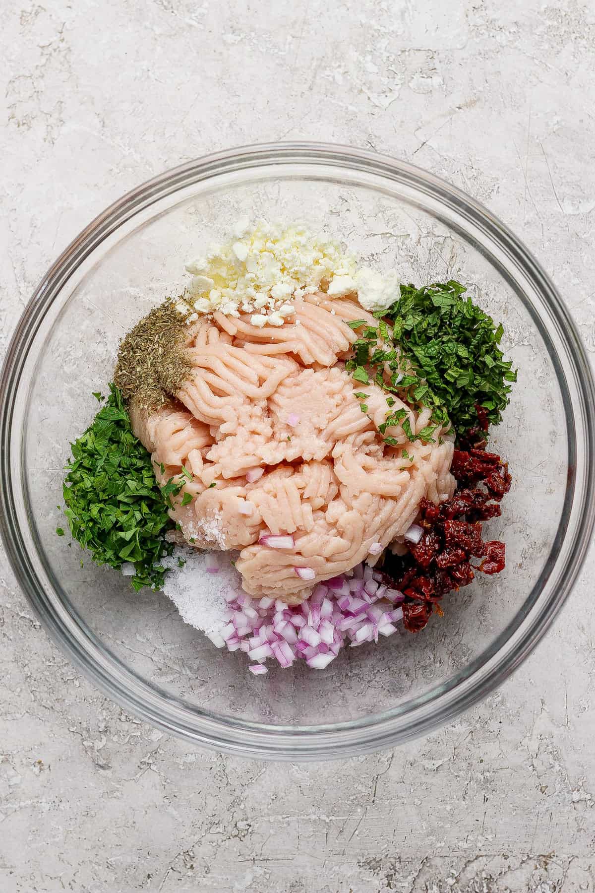 A glass bowl containing ground meat, chopped red onion, herbs, minced garlic, sun-dried tomatoes, salt, pepper, and seasonings arranged separately.