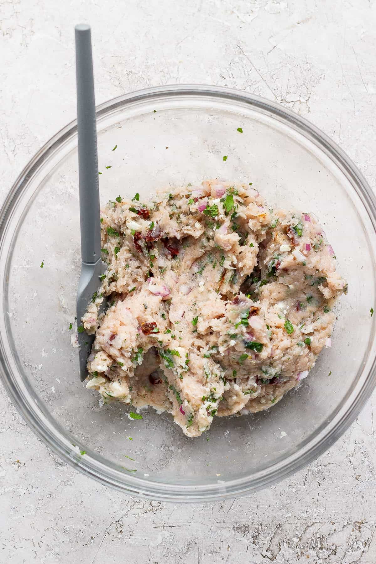 A glass bowl with a gray spoon contains a mixture of mashed white beans, chopped herbs, red onion, and sun-dried tomatoes on a light-colored surface.