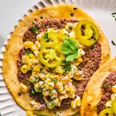 A close-up of a taco with ground beef, creamy corn salad, pickled jalape&ntilde;o slices, and a sprig of cilantro on a white plate.