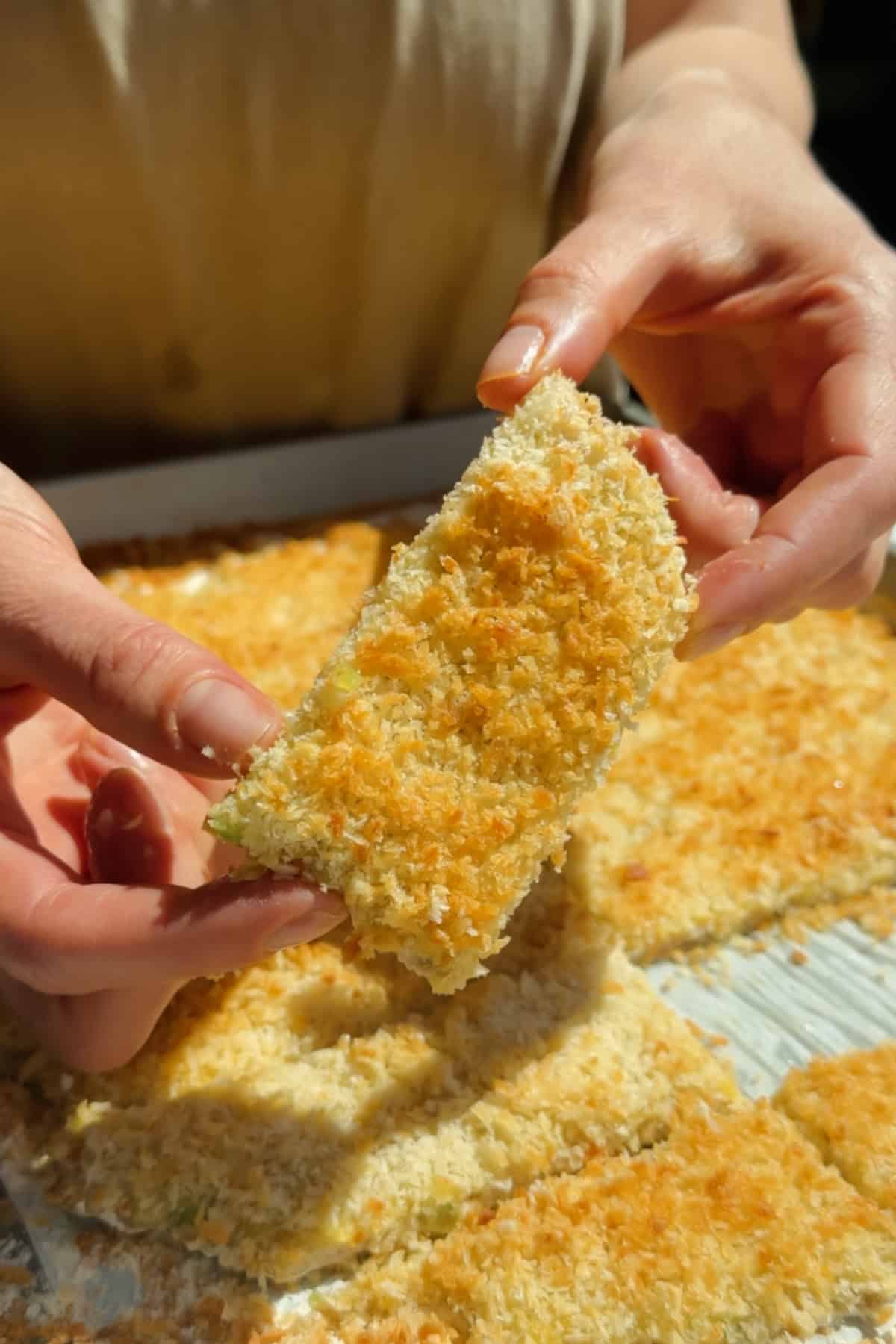 Holding a golden crispy chicken protein bar with panko coating above a tray of baked bars