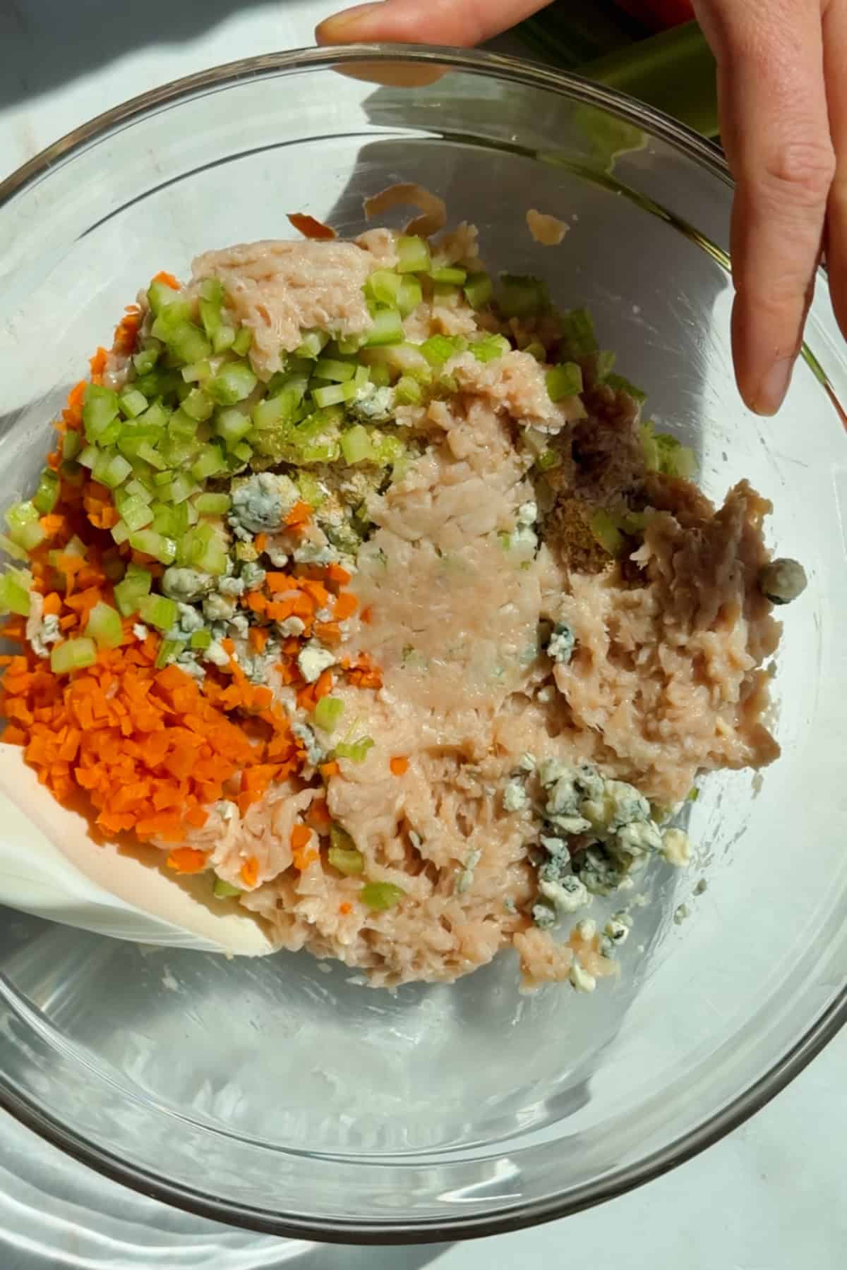 Mixing ground chicken, carrots, celery, blue cheese crumbles, and seasonings in a glass bowl for protein bars