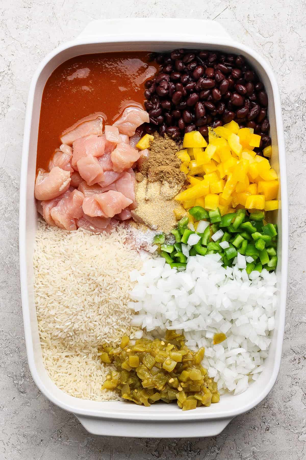 A casserole dish with raw chicken, black beans, enchilada sauce, yellow bell pepper, green bell pepper, chopped onions, green chilies, rice, and seasonings arranged in sections.