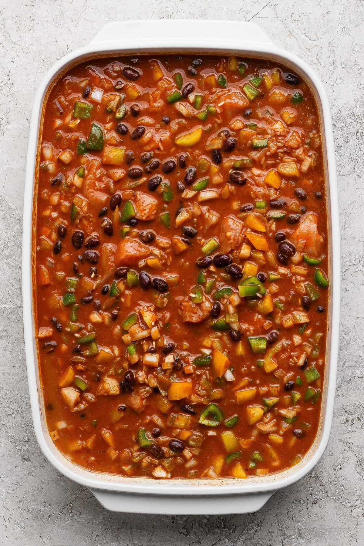 A rectangular white casserole dish filled with a chunky mixture of beans, tomatoes, rice, and vegetables in a red sauce on a textured surface.