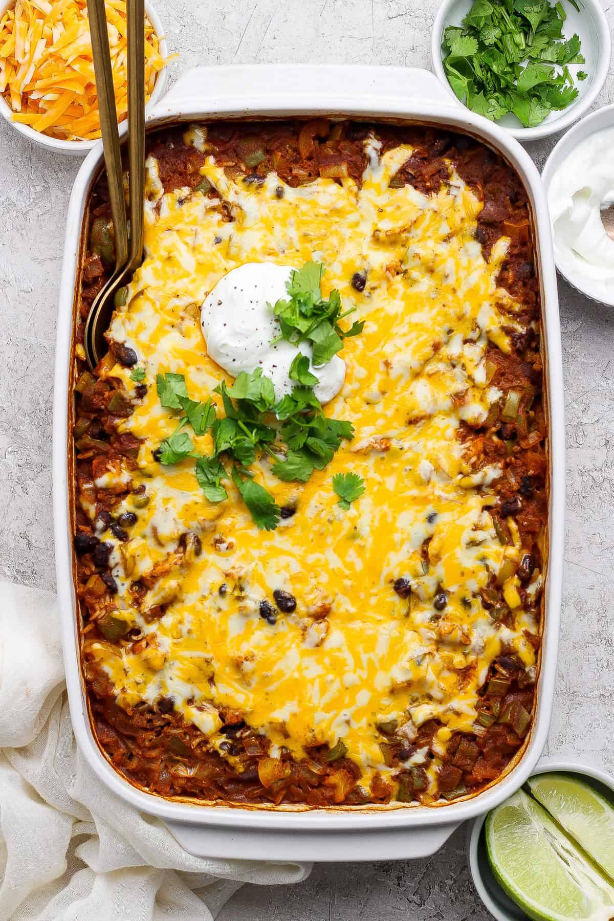 A baked casserole topped with melted cheese, sour cream, and fresh cilantro, in a white dish, with lime wedges, shredded cheese, and sour cream in bowls on the side.