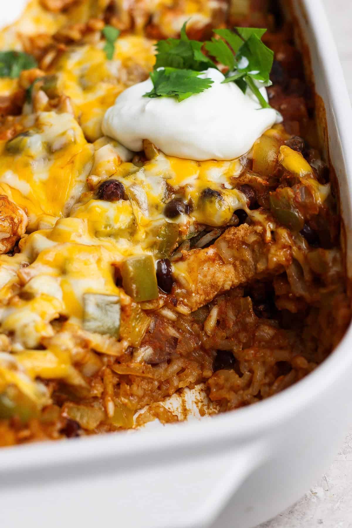 A baked casserole with melted cheese, rice, beans, and green peppers, topped with a dollop of sour cream and fresh cilantro in a white dish.