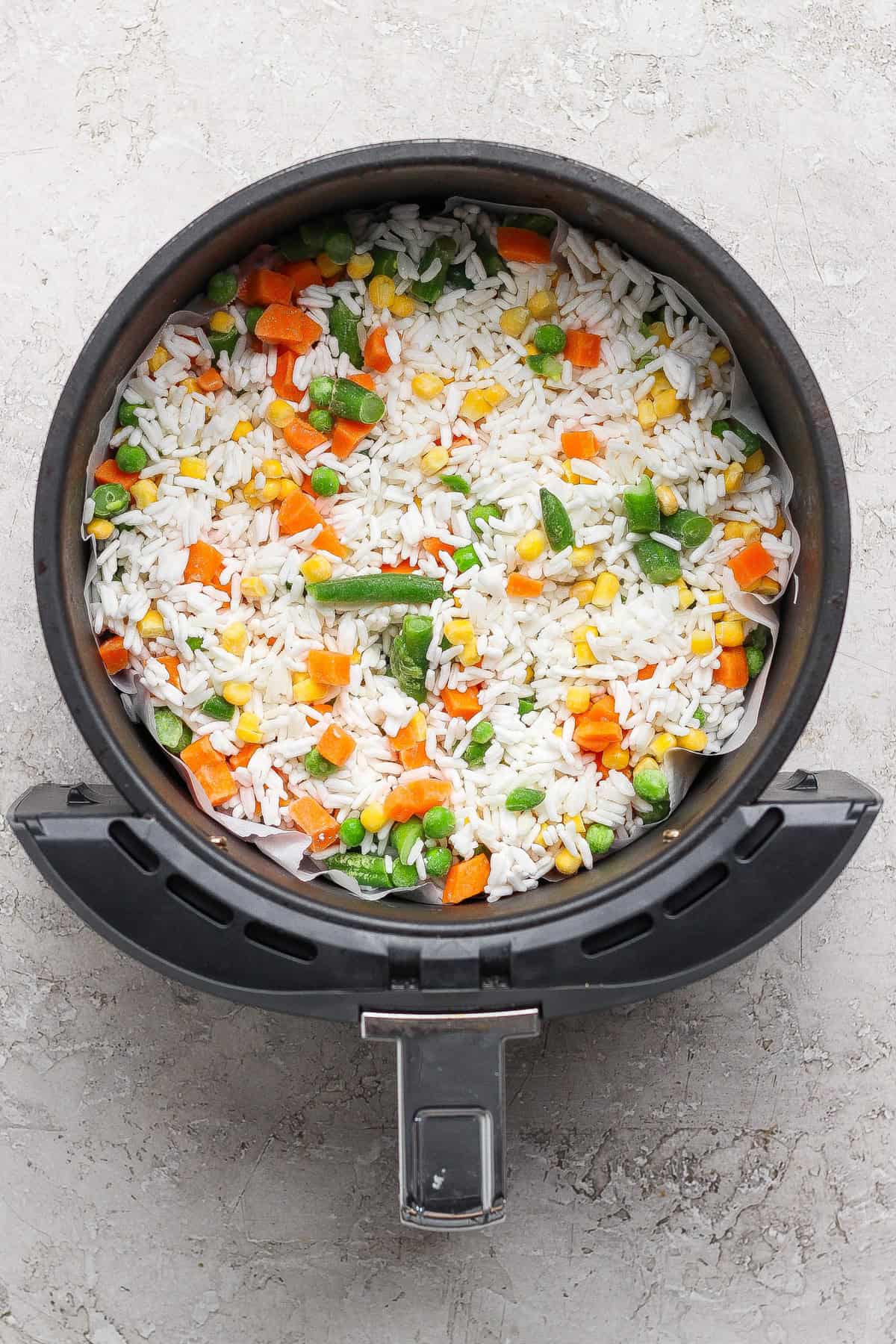 An air fryer basket filled with uncooked white rice and mixed vegetables including carrots, peas, corn, and green beans.