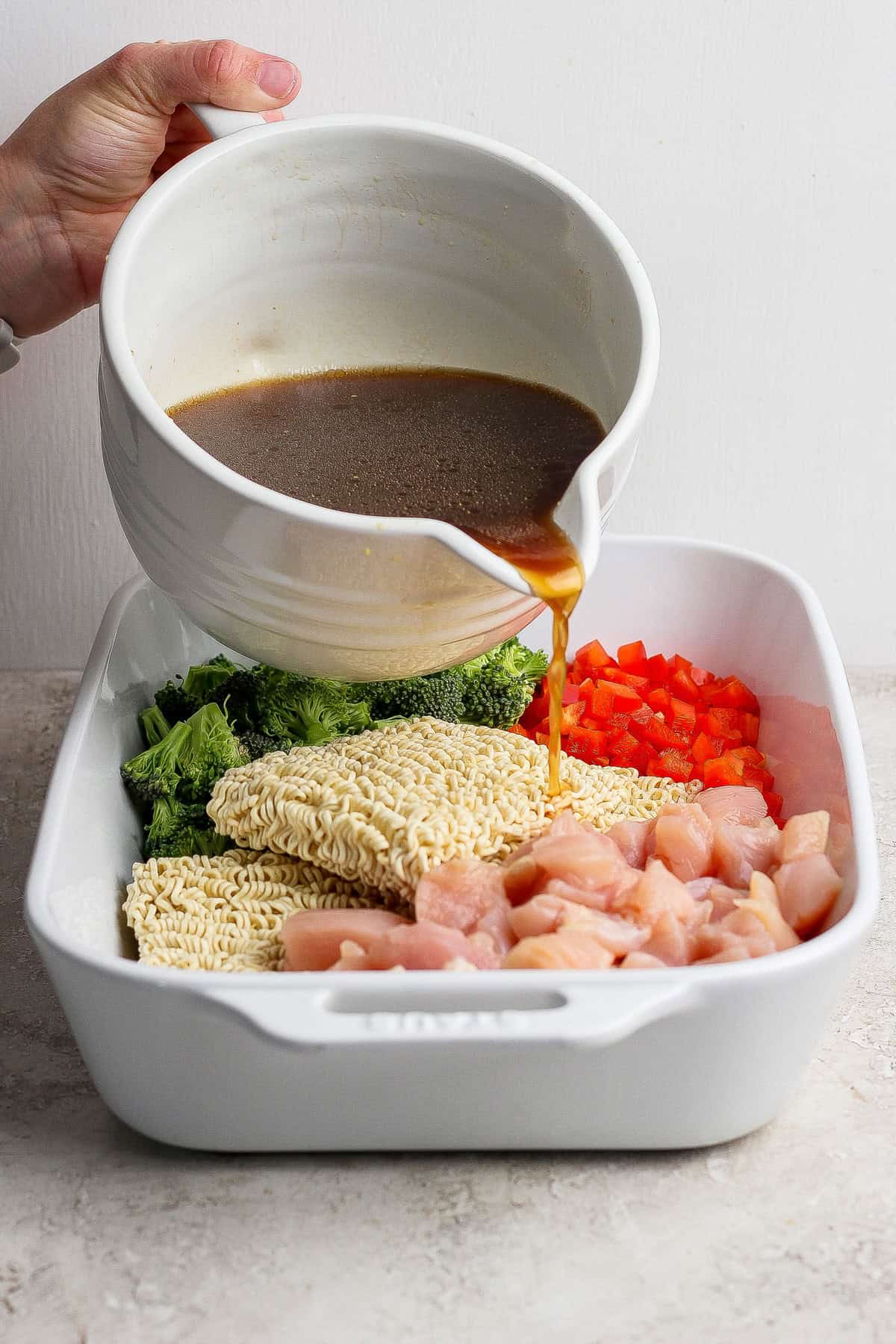 A hand pours brown sauce from a white bowl over uncooked ramen noodles, chopped broccoli, red bell pepper, and diced chicken in a white baking dish.