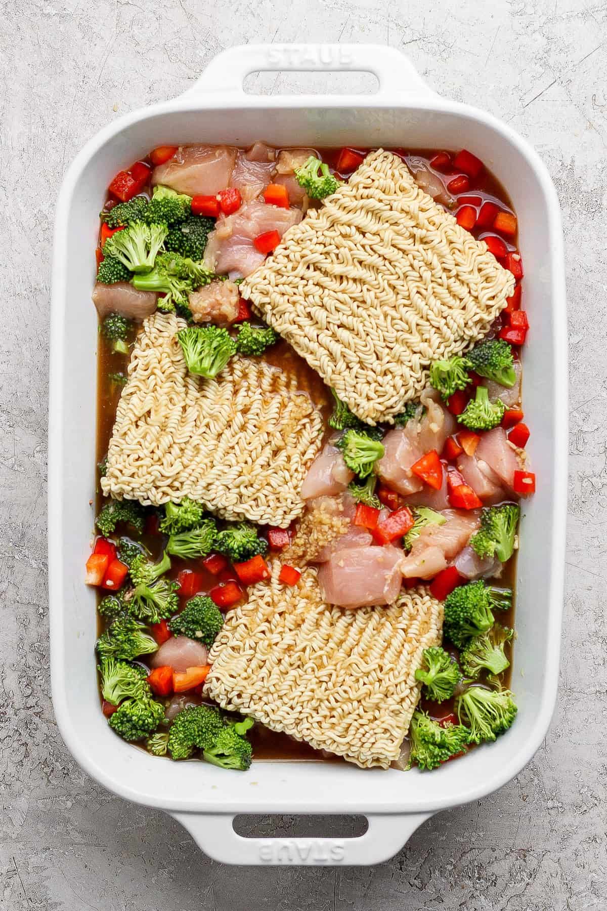 Uncooked ramen noodles, raw chicken pieces, chopped broccoli, and diced red bell peppers in a white rectangular baking dish.