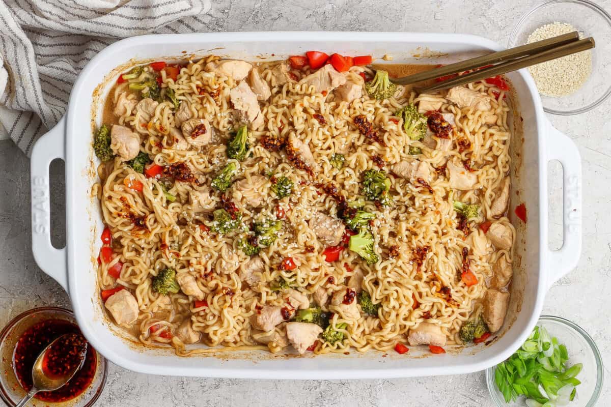Rectangular white baking dish filled with cooked ramen noodles, chicken pieces, broccoli, and red peppers, garnished with sesame seeds and sauce, with chopsticks resting on the dish.