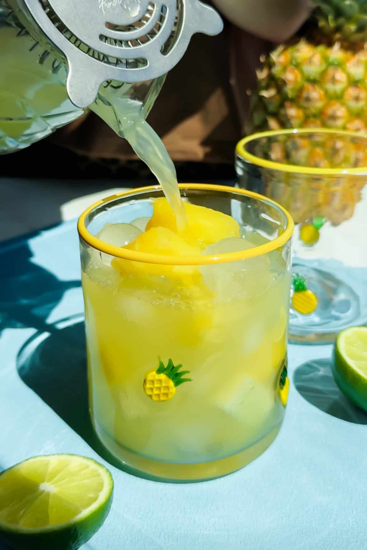 A glass with ice and yellow frozen fruit is being filled with juice. The glass has pineapple designs. Lime halves and another glass are nearby.