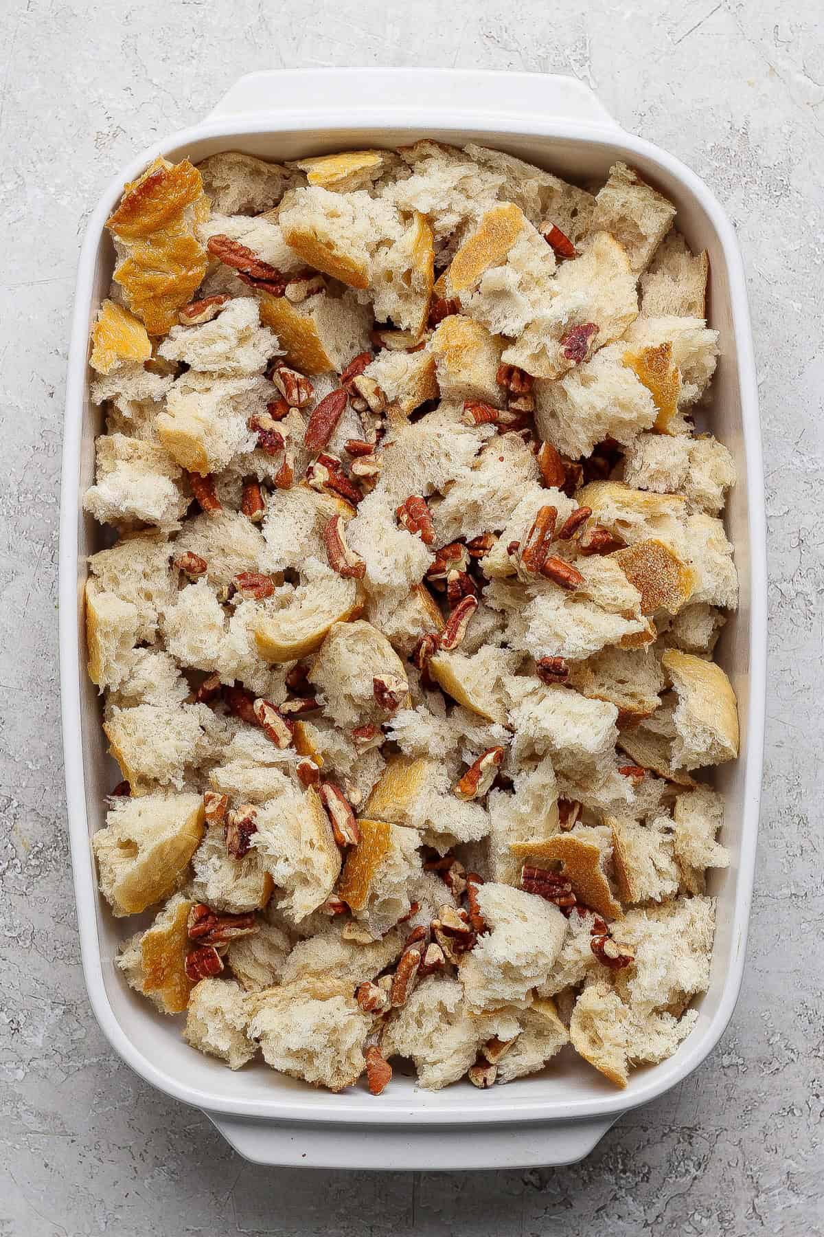 A white baking dish filled with chunks of bread and chopped pecans on a light-colored surface.