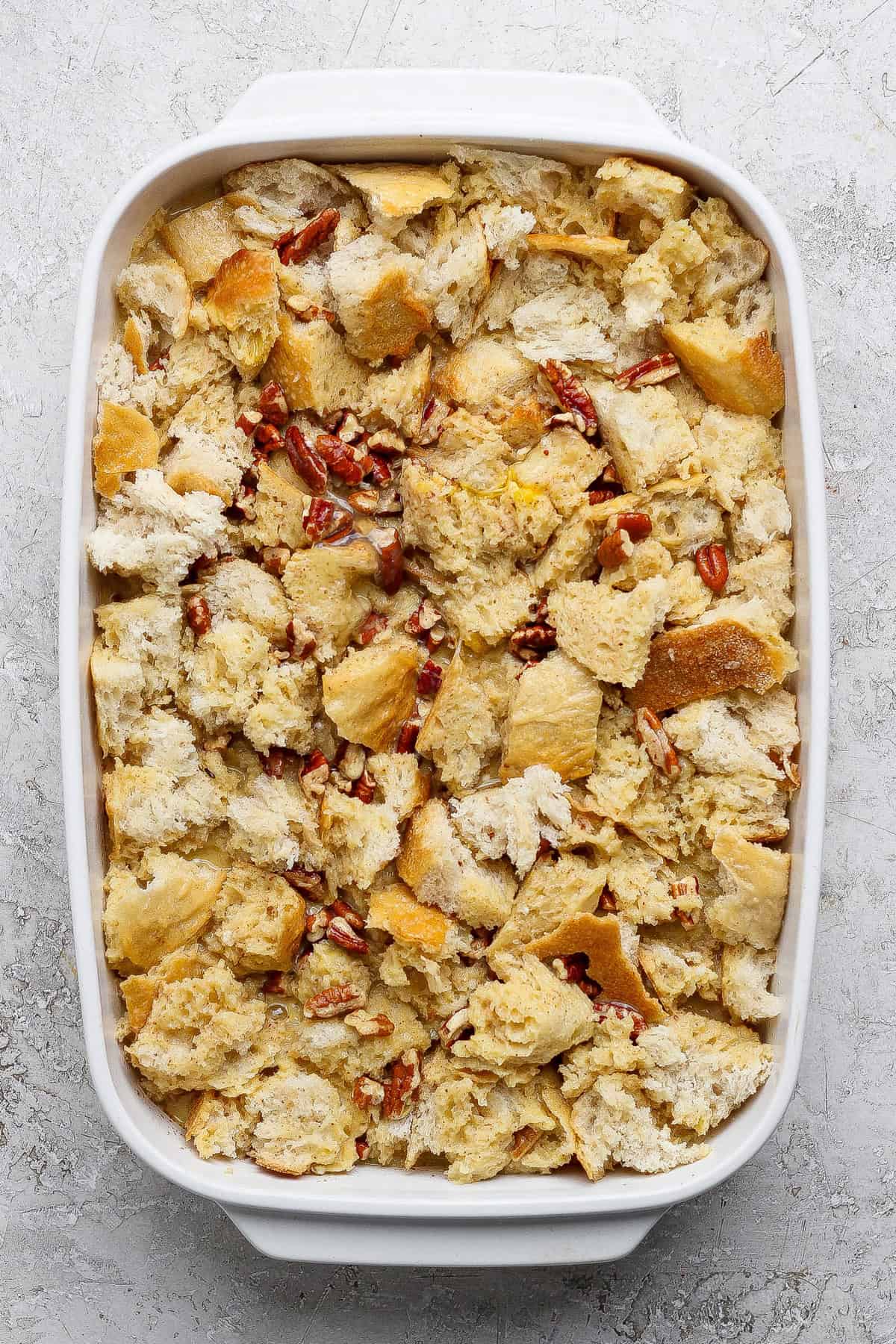 A white baking dish filled with unbaked bread pudding made of torn bread pieces and pecans on a light textured surface.