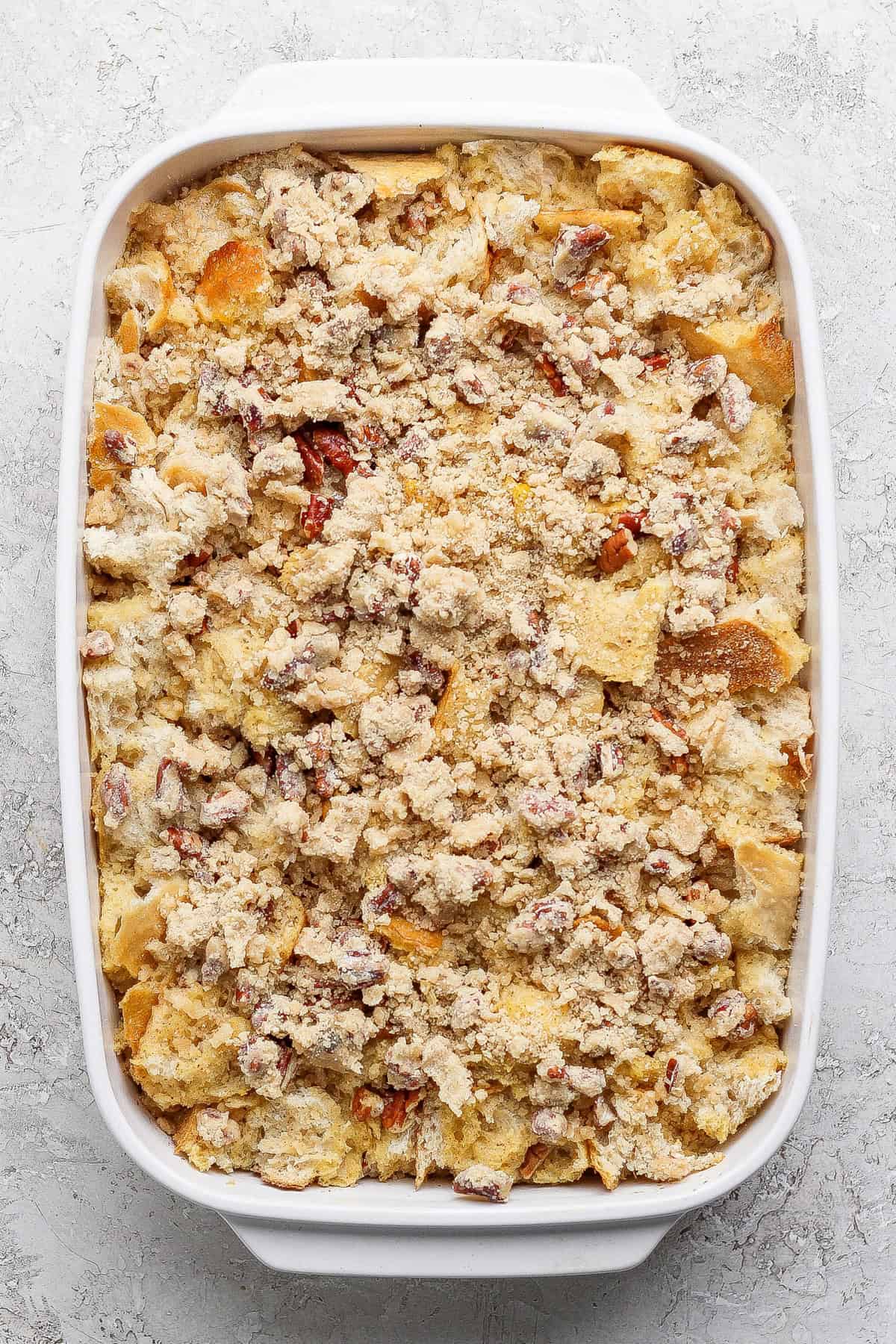 A rectangular white baking dish filled with a crumbly casserole or bread pudding mixture, topped with chopped nuts or streusel, on a light textured surface.