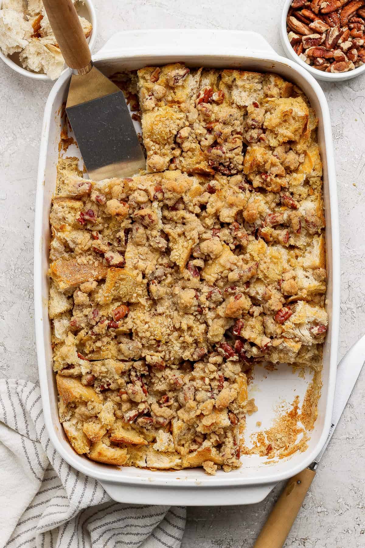 A baked casserole dish filled with pecan-topped bread pudding, partially sliced, with a spatula and a knife nearby. Small bowls of pecans and bread pieces are visible in the background.