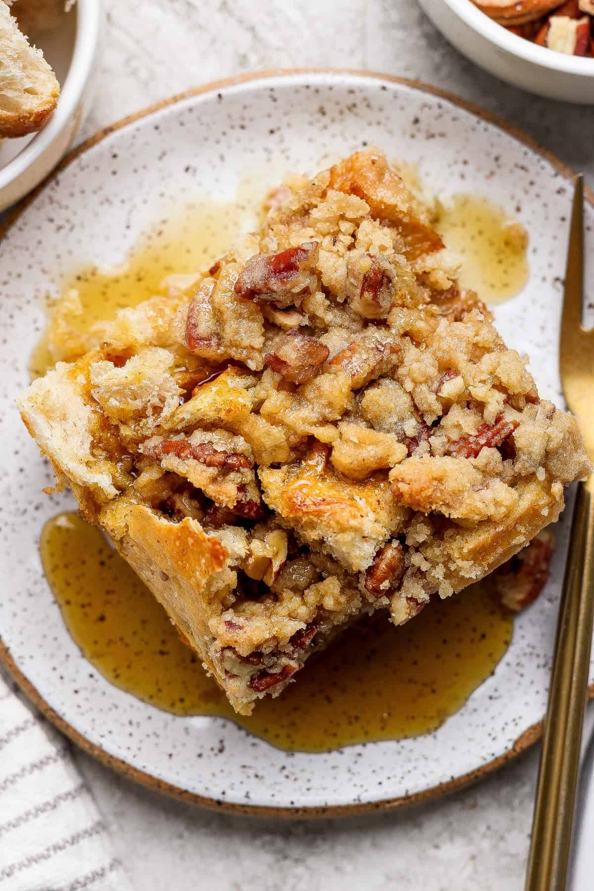 A square slice of pecan-topped bread pudding sits on a speckled plate with syrup drizzled around it and a gold fork nearby.
