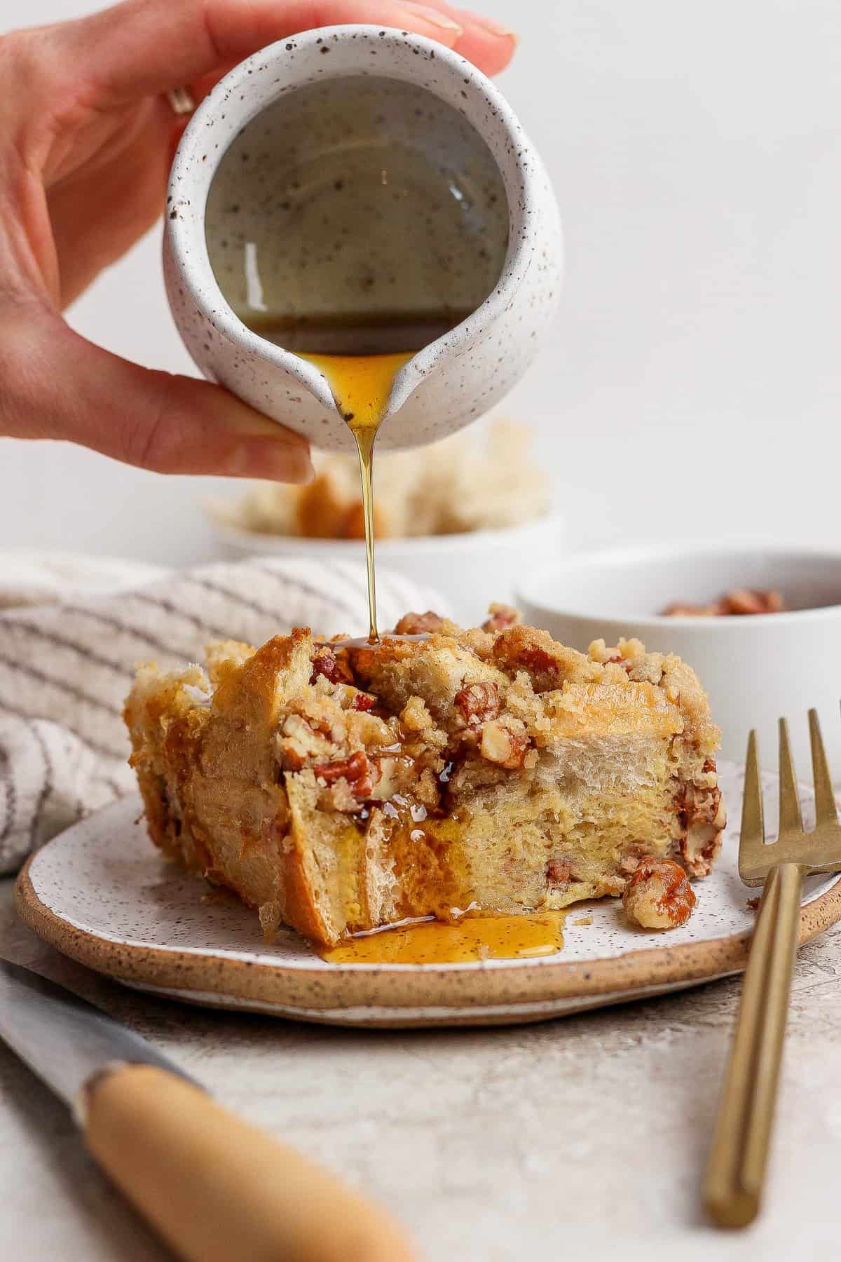 A hand pours syrup from a small pitcher onto a slice of bread pudding topped with nuts on a plate, with a fork and knife beside it.